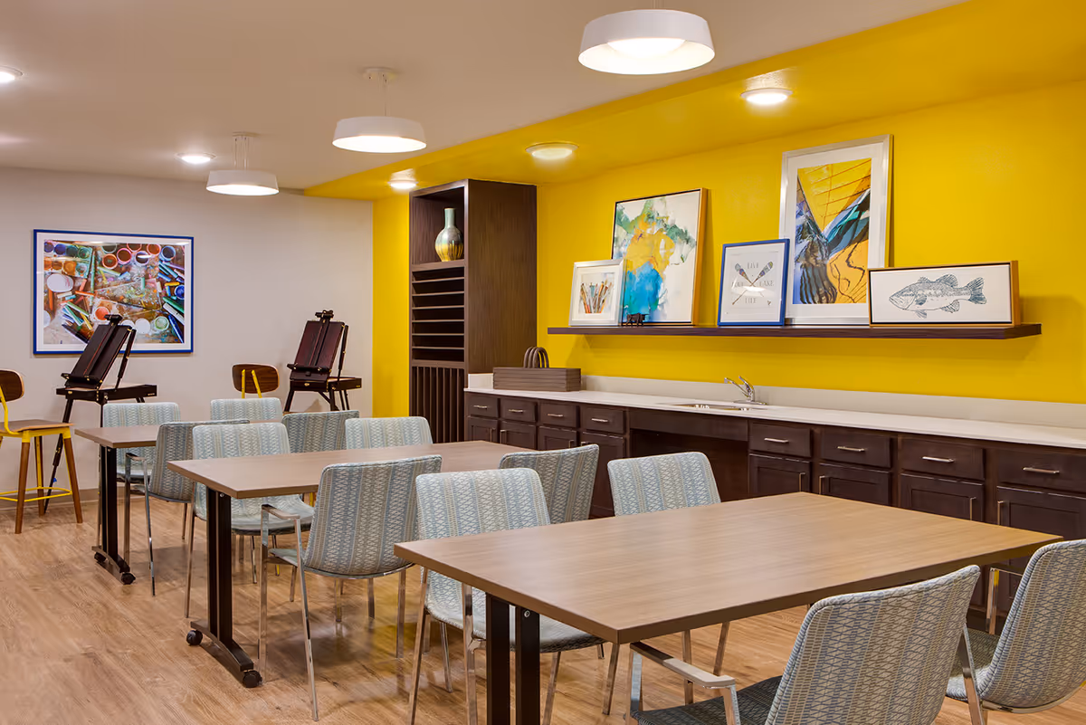 A bright and colorful activity room with wooden tables and patterned chairs arranged for group seating. The back wall is painted yellow and decorated with framed artwork and a shelf. There are cabinets and a countertop with a sink beneath the artwork. In the background, there are easels and a framed picture on a white wall. The room has wood flooring and modern ceiling lights.
