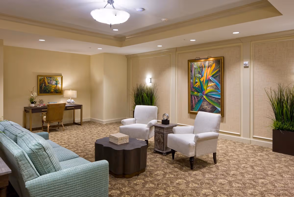 A cozy living room area with a light blue sofa, two white armchairs, a dark wooden coffee table, and a small side table. The walls are beige with decorative paneling, featuring two colorful paintings and two tall green plants. There is a desk with a chair and a lamp in the corner, and the room is softly lit by ceiling lights and a central chandelier.