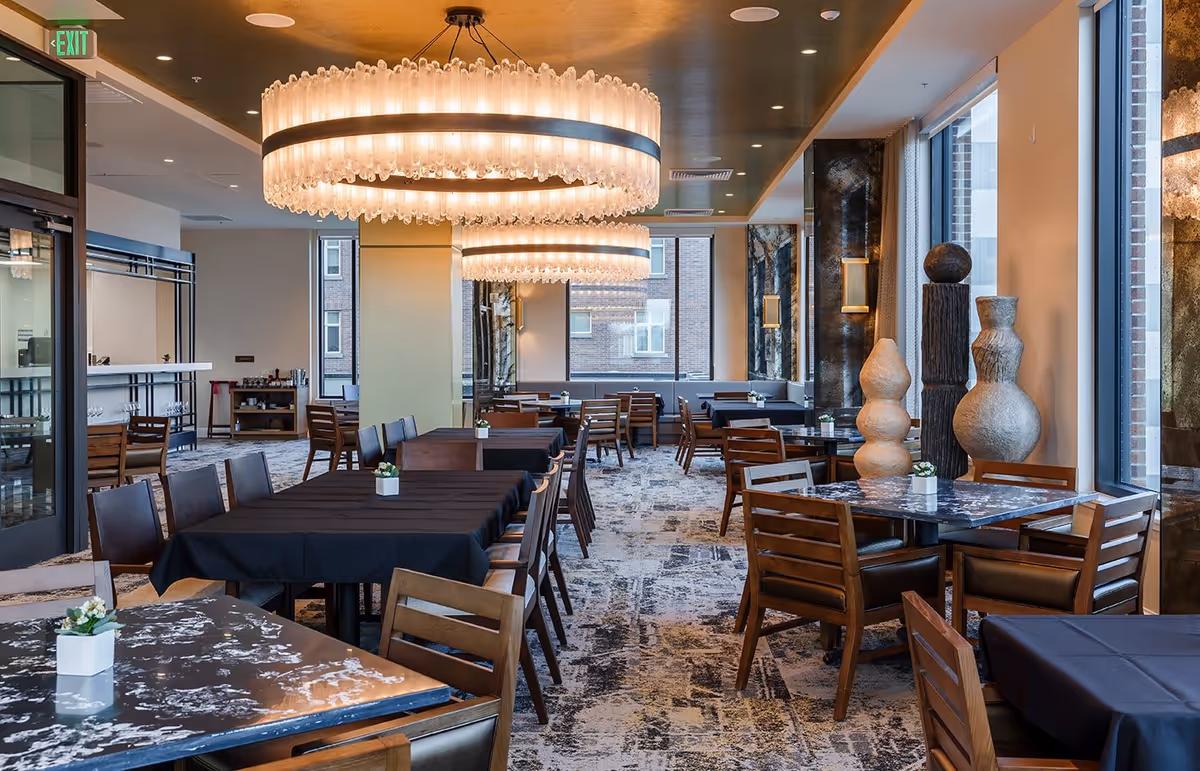 A modern dining room with multiple tables covered in black tablecloths and marble-patterned tabletops, surrounded by wooden chairs. Large circular chandeliers hang from the ceiling, and decorative sculptures are placed near the windows. The room has large windows letting in natural light and a patterned carpet floor.