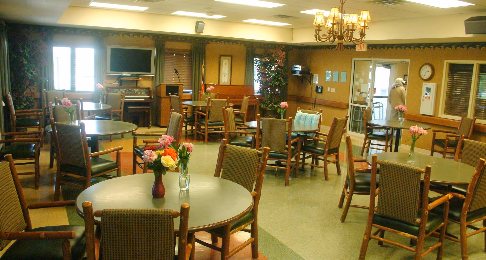 A well-lit common area with multiple round tables and wooden chairs arranged around them. Each table has a small vase with colorful flowers. The room has a chandelier, a piano, a television mounted on the wall, and large windows with blinds. There is a person near the exit door in the background.