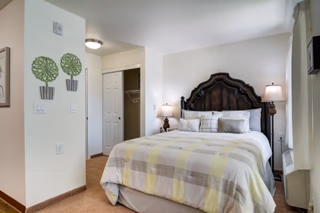 A cozy bedroom with a large bed featuring a dark wooden headboard and a light-colored plaid bedspread. There are two bedside tables with lamps on each side of the bed. The room has beige walls, a carpeted floor, a closet with white sliding doors, and decorative green wall art on one wall.