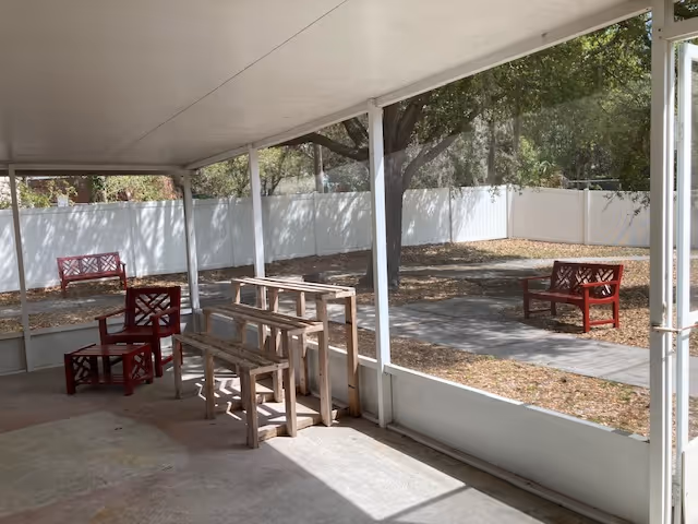 Covered outdoor patio with wooden benches, red chairs, and a white fence surrounding trees.
