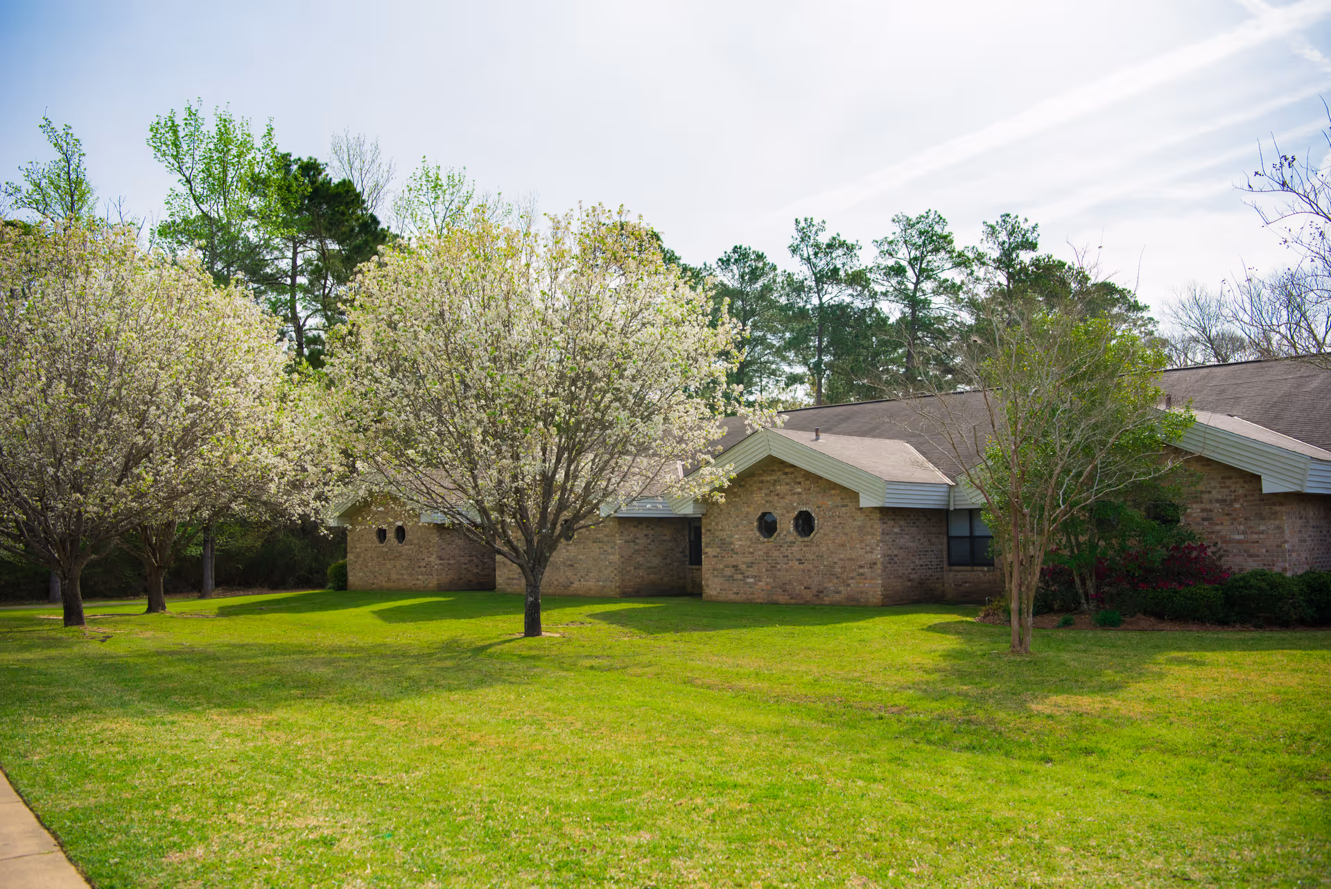 A single-story brick building with a sloped roof surrounded by green grass and several blossoming trees. The sky is partly cloudy, and there are taller trees in the background.