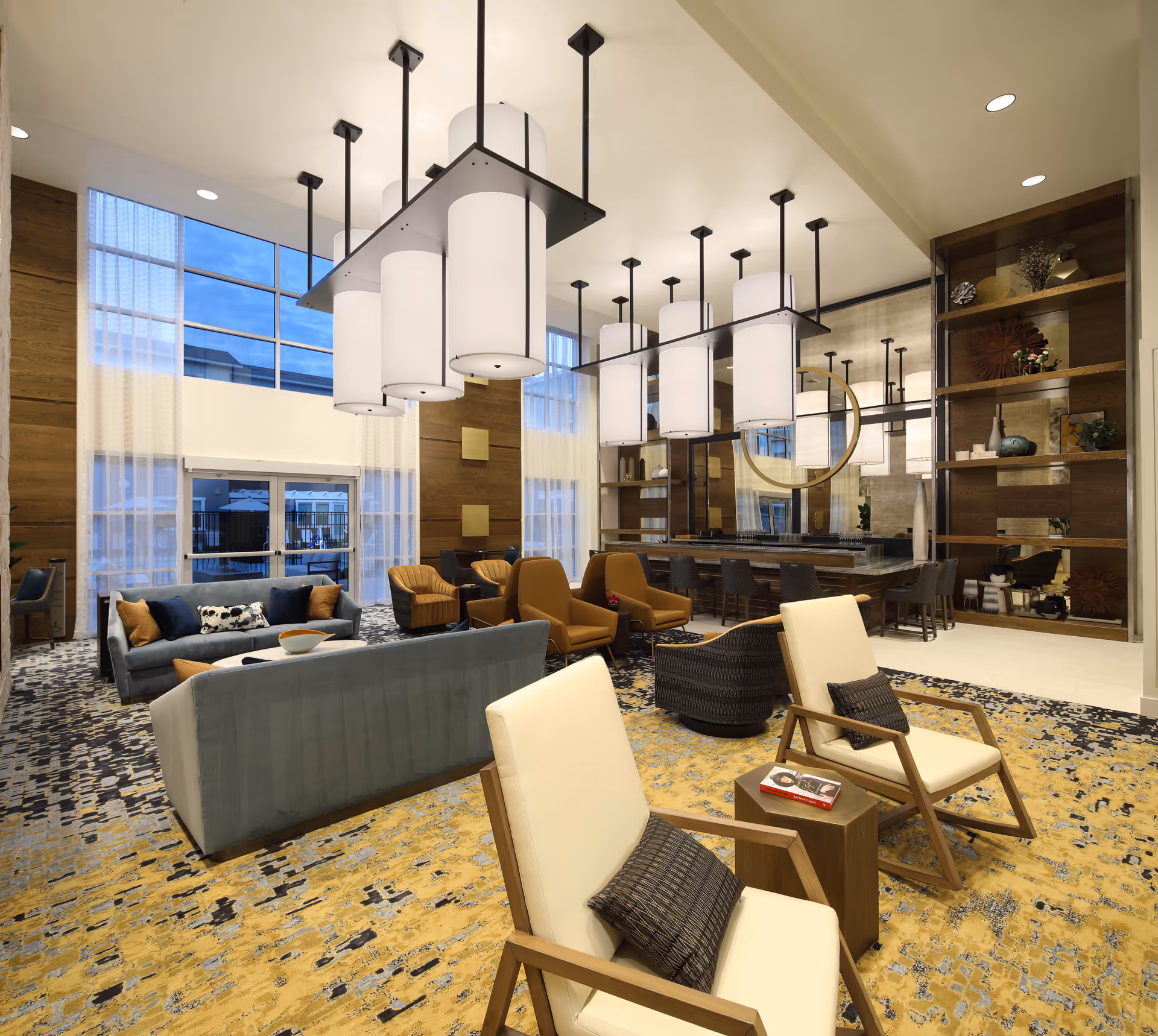 A spacious and modern senior living common area with high ceilings and large windows. The room features multiple seating arrangements including cream-colored armchairs with dark cushions, blue sofas, and brown armchairs. There are large cylindrical pendant lights hanging from the ceiling, a patterned carpet in shades of yellow, blue, and gray, and a decorative shelving unit with various ornaments. A large mirror with a circular frame is mounted on the wall behind a bar-height counter with stools.