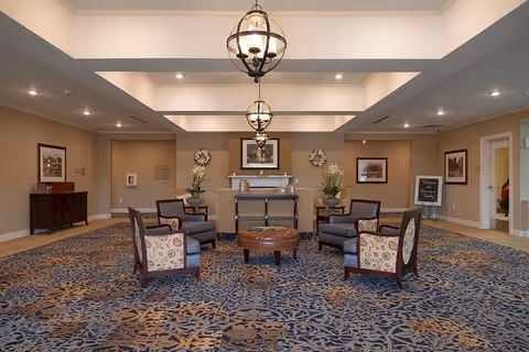 A spacious and elegant common area with a patterned carpet, four upholstered armchairs arranged around a round ottoman, two large potted plants, framed artwork on the walls, and two hanging pendant lights from a coffered ceiling.