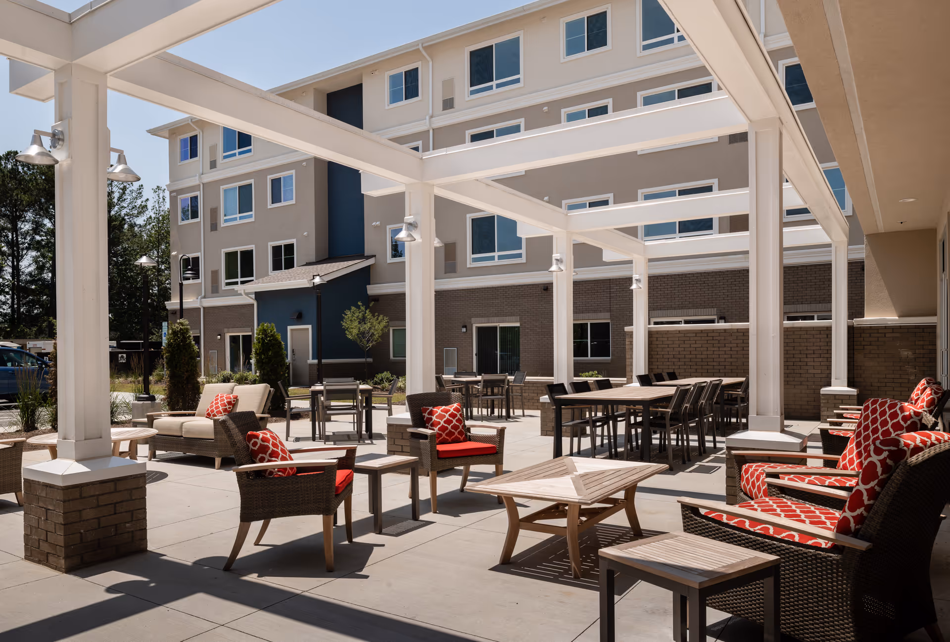 Outdoor patio area at The Willows at Raleigh featuring multiple seating arrangements with cushioned chairs and sofas in red and beige tones, wooden tables, and white pergola structures. The background shows a multi-story building with numerous windows and a mix of beige, blue, and brick exterior walls.
