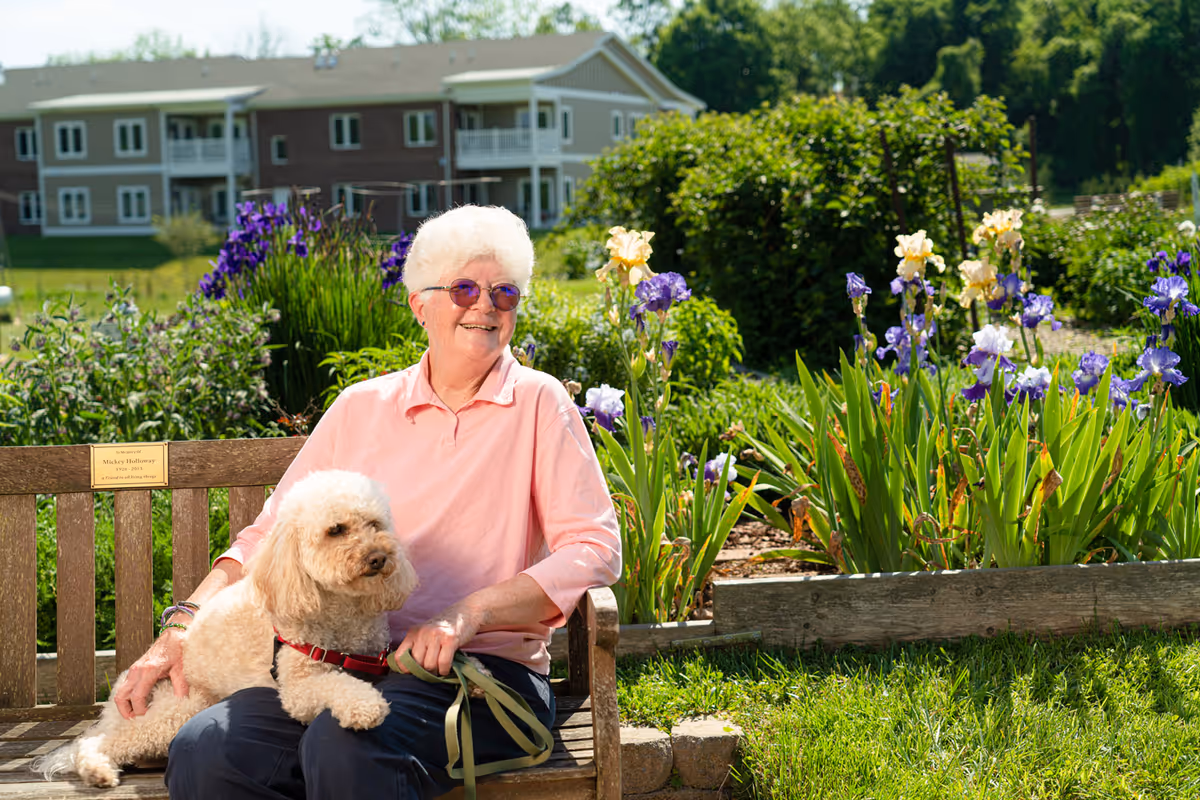 An elderly woman with short white hair and purple sunglasses sits on a wooden bench in a garden, smiling while holding a small fluffy dog on her lap. Behind her are colorful flowers and greenery, with a large residential building in the background under a clear sky.