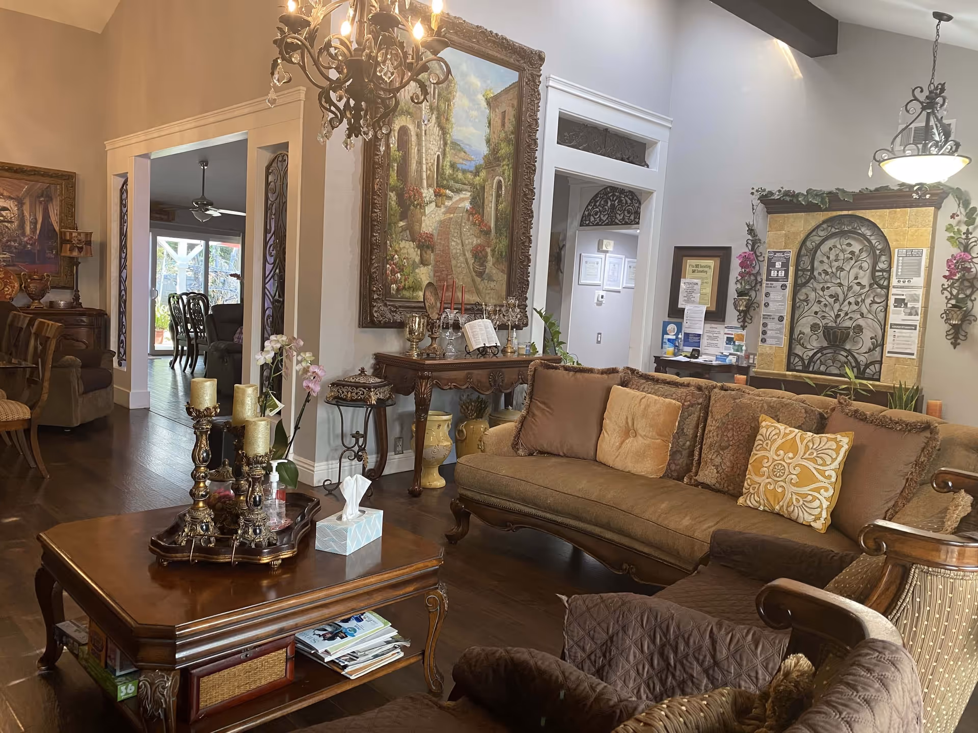 A cozy living room with a brown sofa adorned with multiple decorative pillows, a wooden coffee table with candles and a tissue box, and an ornate chandelier hanging from the ceiling. The room features elegant wall art, decorative plants, and a warm, inviting atmosphere with wooden flooring and classic furniture.