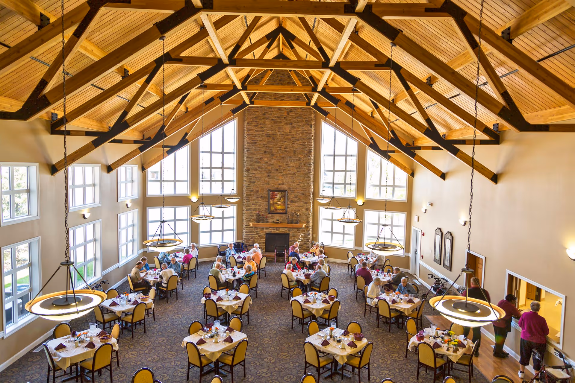 A spacious dining room with high vaulted wooden ceilings and large windows letting in natural light. Several round tables are set with tablecloths, napkins, and dining ware. Elderly people are seated at the tables, eating and socializing. There is a stone fireplace on the far wall with a framed picture above it. Pendant lights hang from the ceiling, illuminating the room.