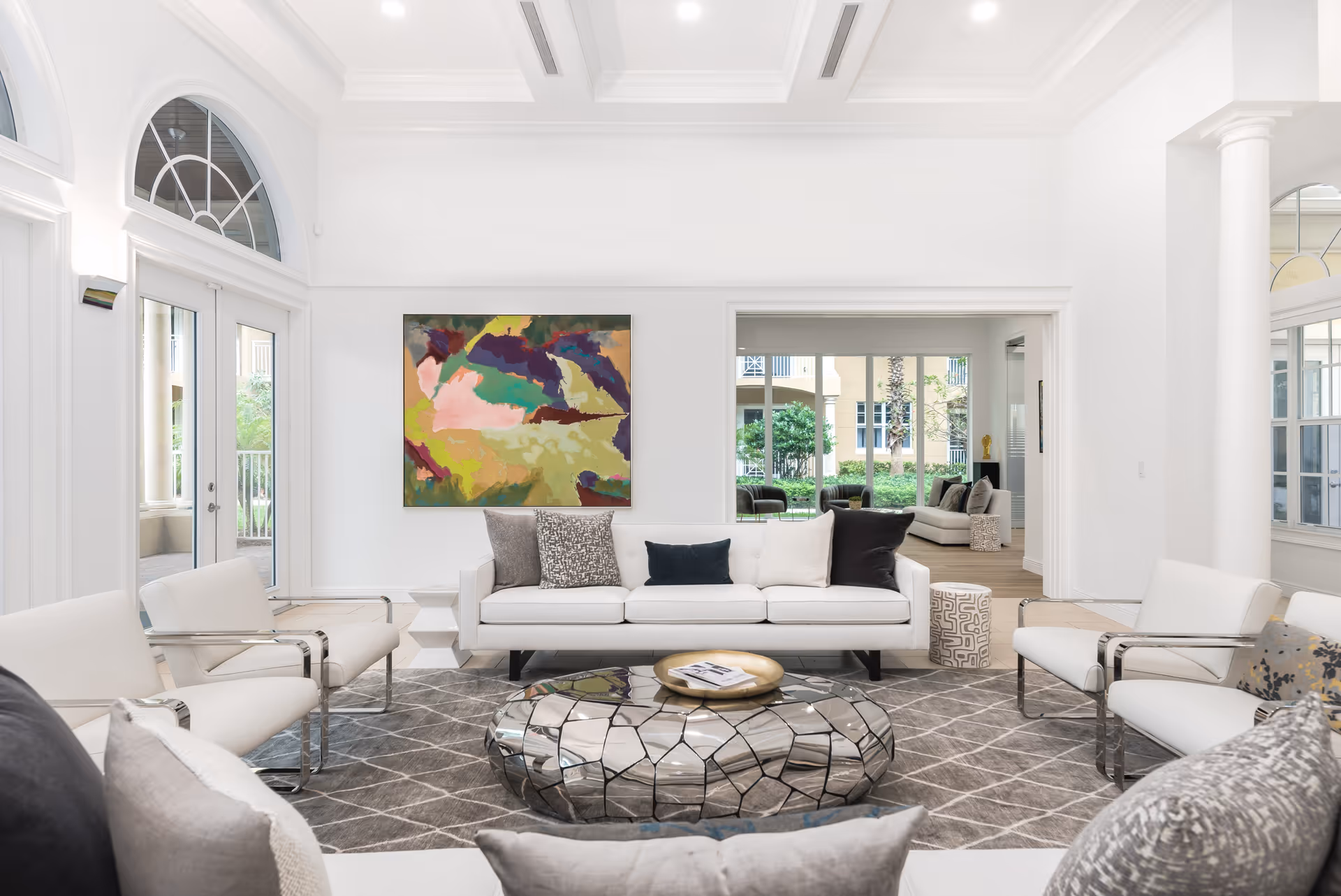 Bright and spacious living room with white walls and high ceilings featuring coffered design. The room has a white sofa with multiple cushions, two white armchairs with chrome frames, and a unique reflective coffee table with a cracked mirror design. A colorful abstract painting hangs on the wall above the sofa. Large windows and glass doors allow natural light to fill the space, and a view into another seating area with similar decor is visible through an open doorway.