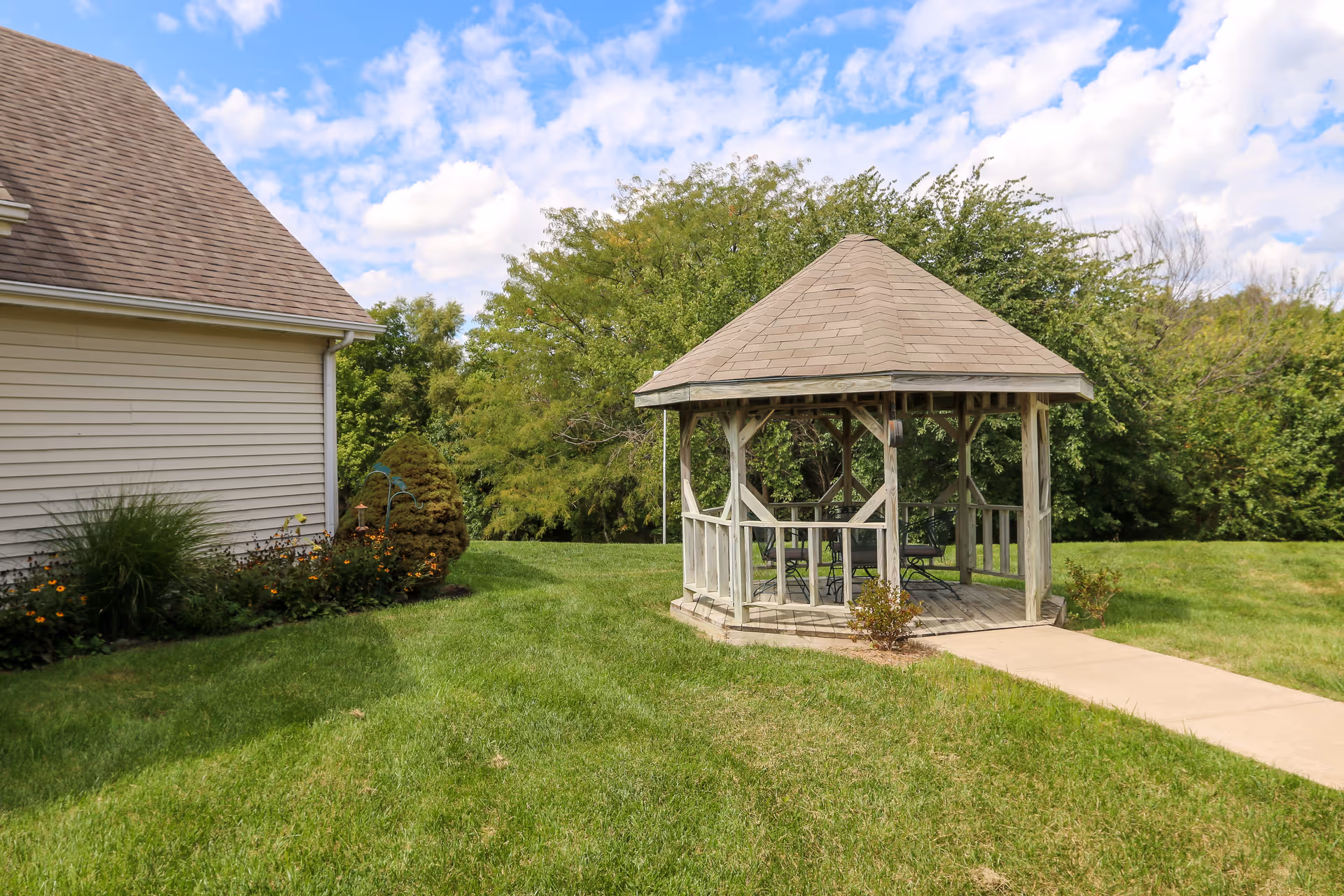 Wooden gazebo on a grassy lawn next to a beige building with trees and a blue sky in the background.