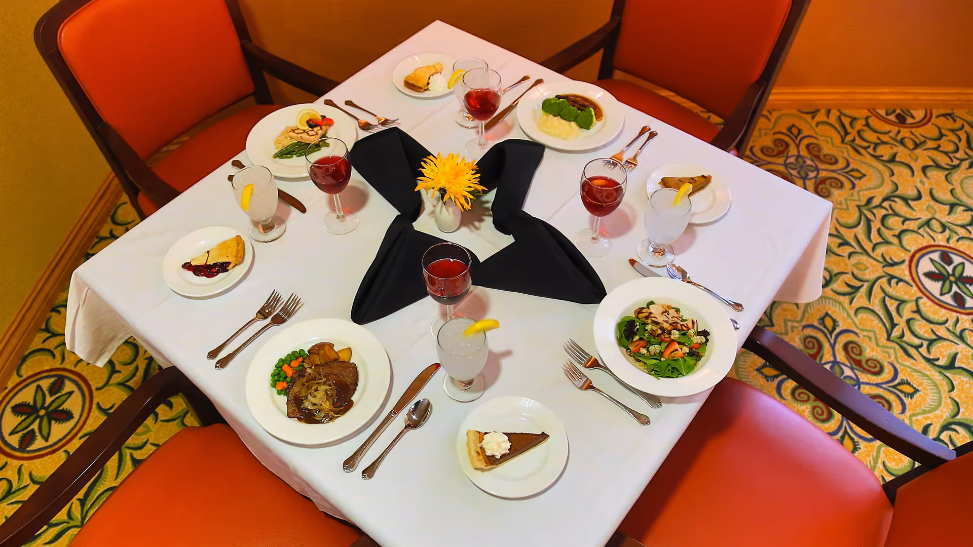 A dining table set for six with white tablecloth, black folded napkins, and a small yellow flower centerpiece. Each place setting includes a plate with a meal such as salad, meat with vegetables, or pie, along with a glass of water with lemon or a glass of red beverage. The table is surrounded by orange cushioned chairs and the floor has a patterned carpet.