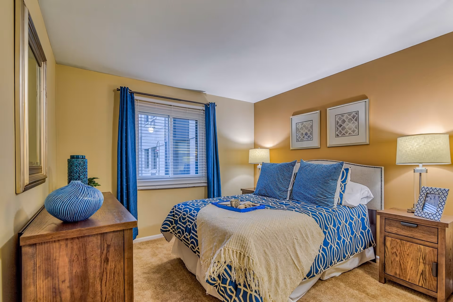 A cozy bedroom with a bed dressed in blue and white patterned bedding and a beige throw blanket. Two blue pillows and two white pillows are arranged on the bed. On either side of the bed are wooden nightstands with lamps and a framed photo on the right nightstand. A wooden dresser with decorative blue vases is on the left side of the room. The walls are painted in a warm beige tone, and there is a window with blue curtains letting in natural light. Two framed artworks hang above the bed.