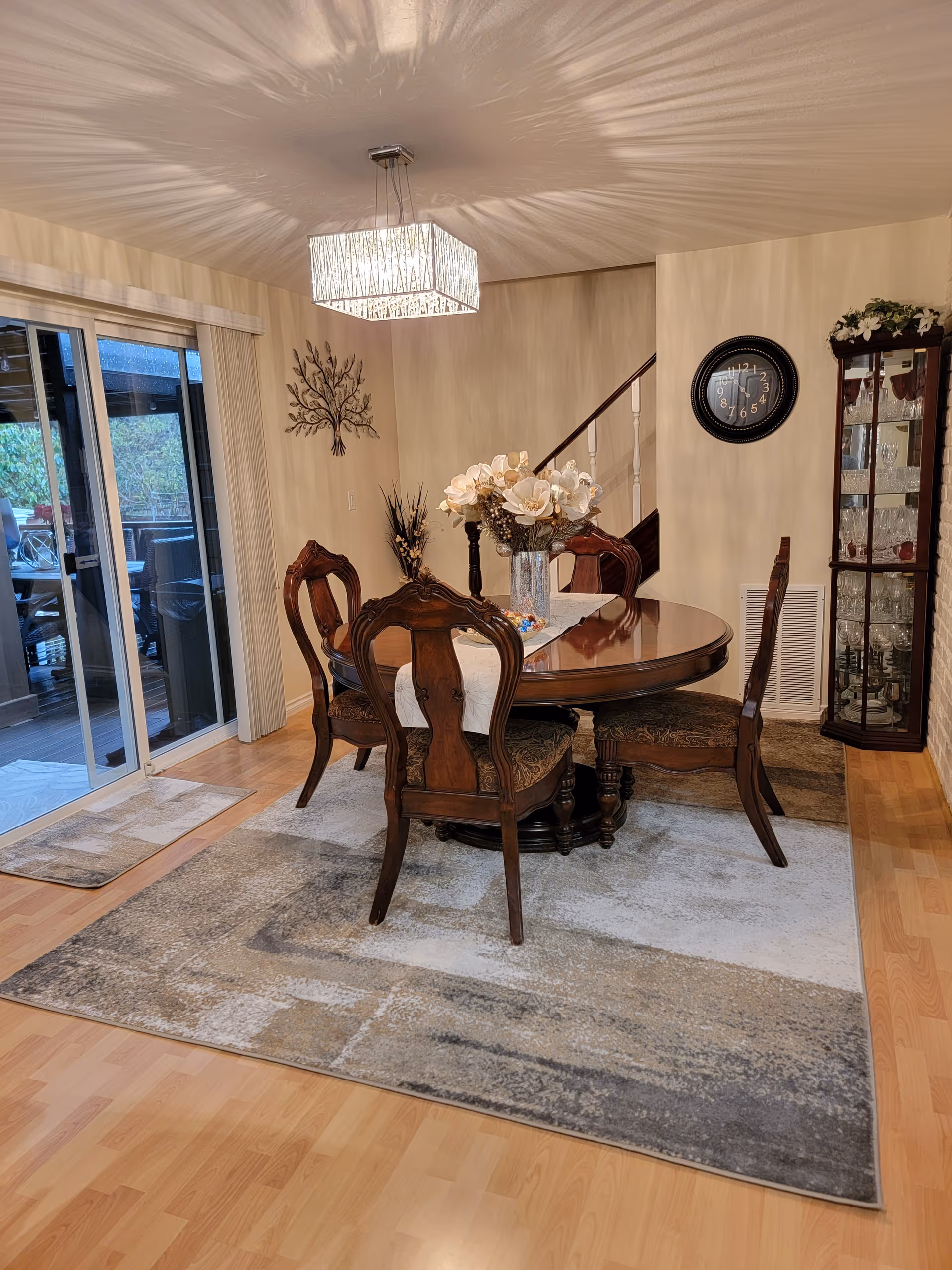 Dining room with a round wooden table and ornate chairs on an area rug beneath a decorative chandelier, sliding glass doors to the left and a china cabinet to the right.