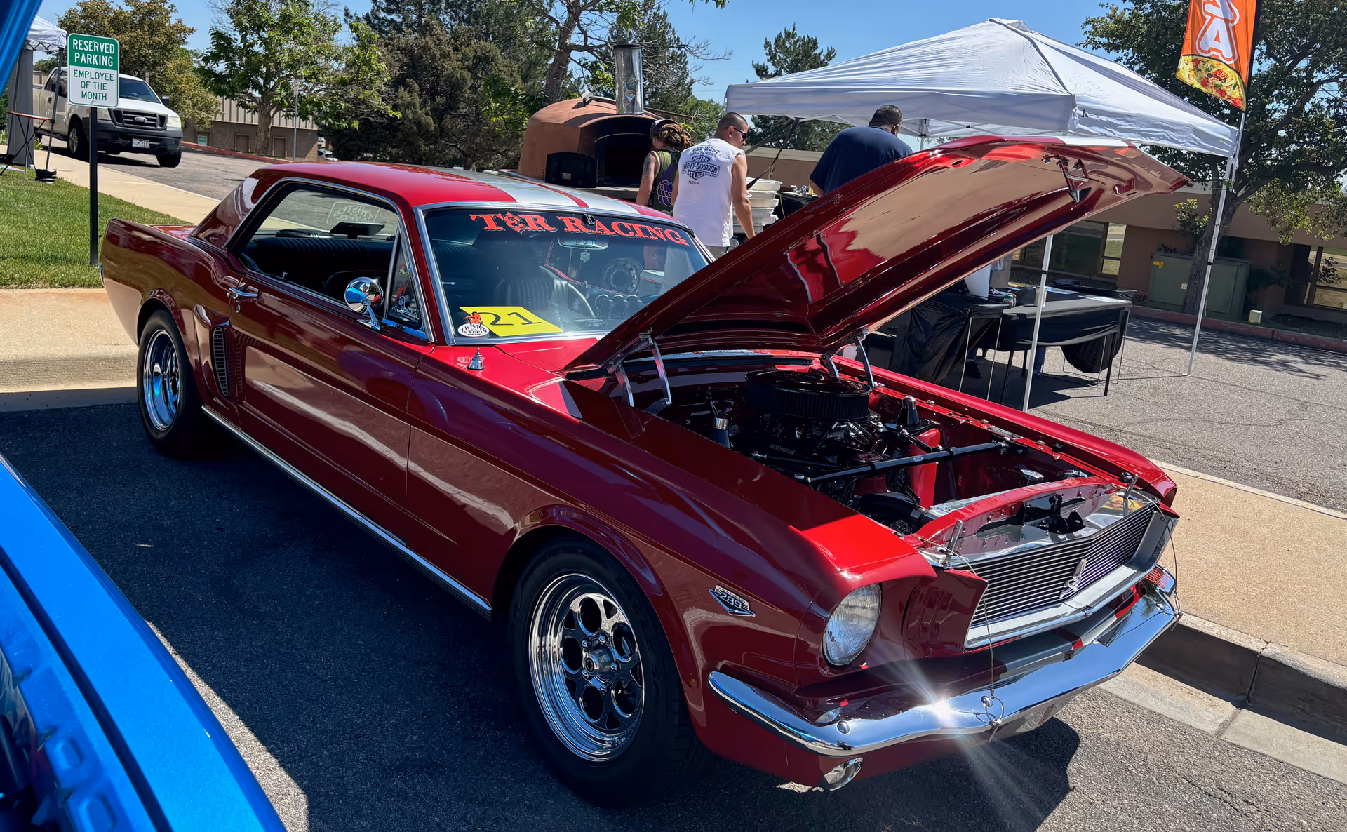 A shiny red classic car with its hood open is parked outdoors on a sunny day. Behind the car, there are a few people standing near a white canopy tent and a pizza oven. A sign indicating reserved parking for employee of the month is visible on the left side of the image.
