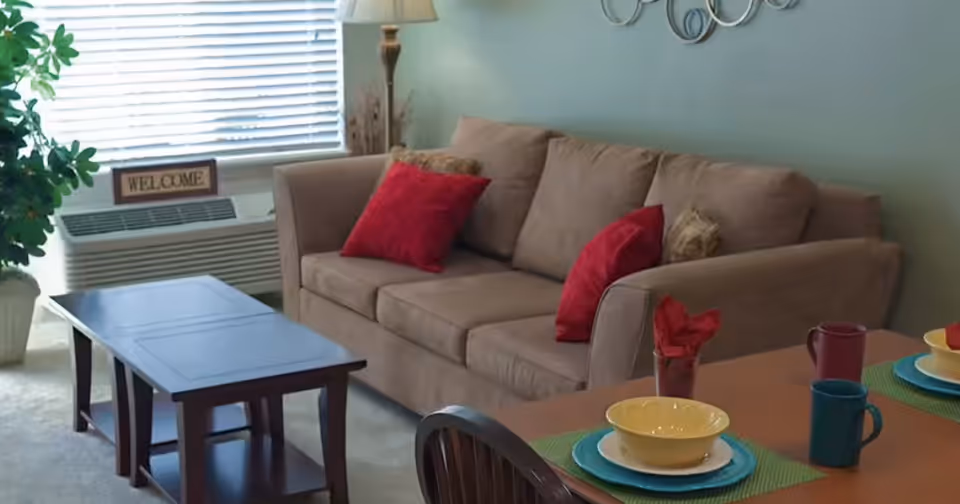 A cozy living room area with a beige sofa adorned with red and patterned throw pillows. In front of the sofa is a dark wooden coffee table. To the left, there is a large green plant in a white pot and a window with blinds partially open, with a small 'WELCOME' sign on the windowsill. On the right side, part of a dining table is visible with colorful place settings including bowls, plates, mugs, and red napkins.