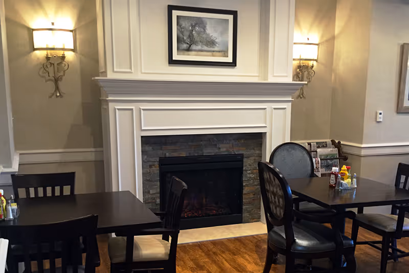 A cozy dining area with dark wooden tables and chairs arranged around an electric fireplace with a white mantel and stone surround. Two wall sconces with lampshades flank the fireplace, and a framed picture hangs above it. Condiments including ketchup and mustard are placed on the tables.