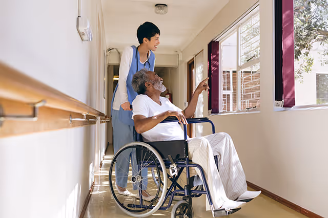 A healthcare worker stands behind an elderly man in a wheelchair in a hallway. The man is pointing out of a window, and both are smiling. The hallway has handrails along the walls and windows letting in natural light.