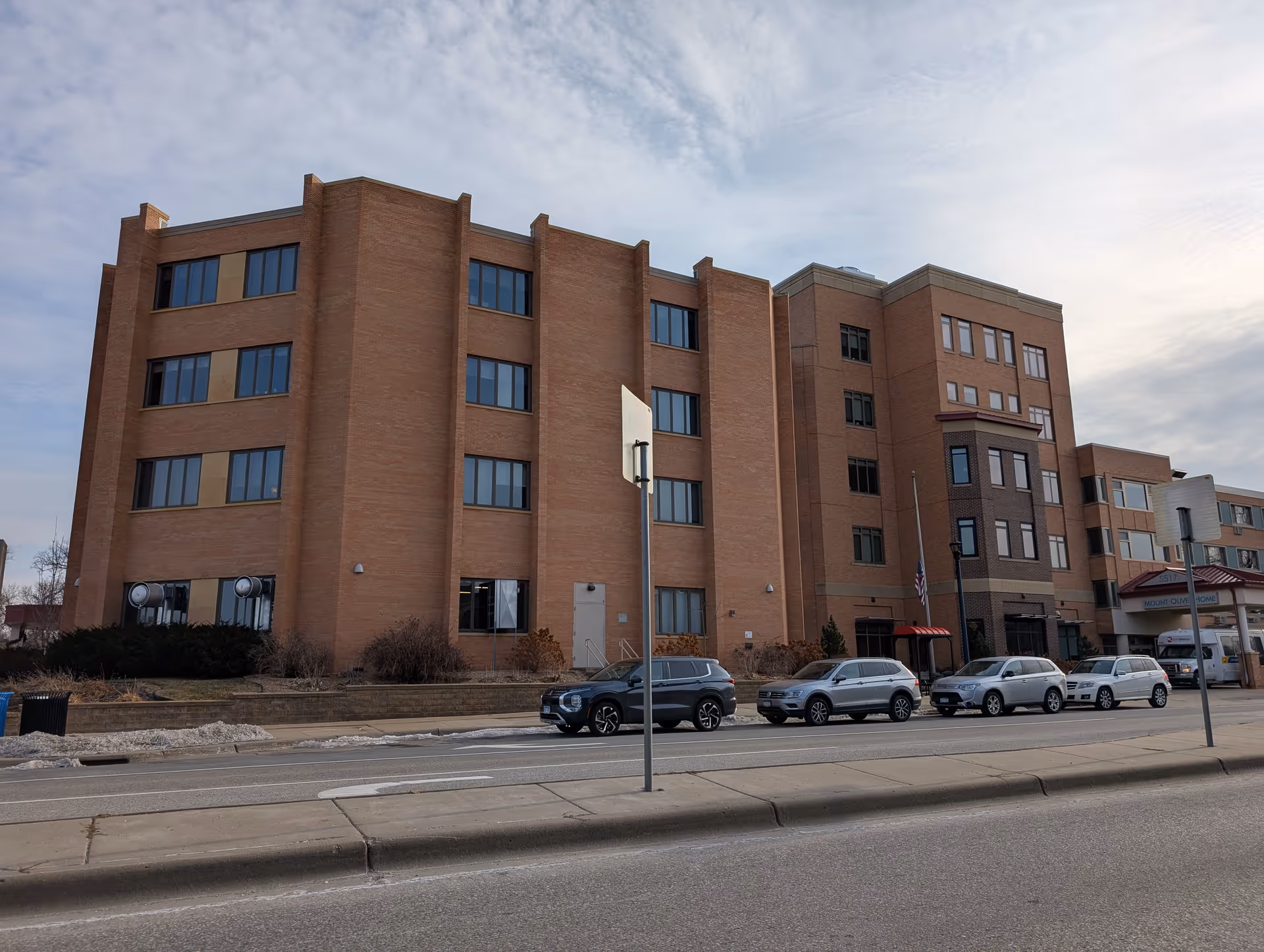 Front exterior of a multi-story brick care facility with several cars parked along the street.