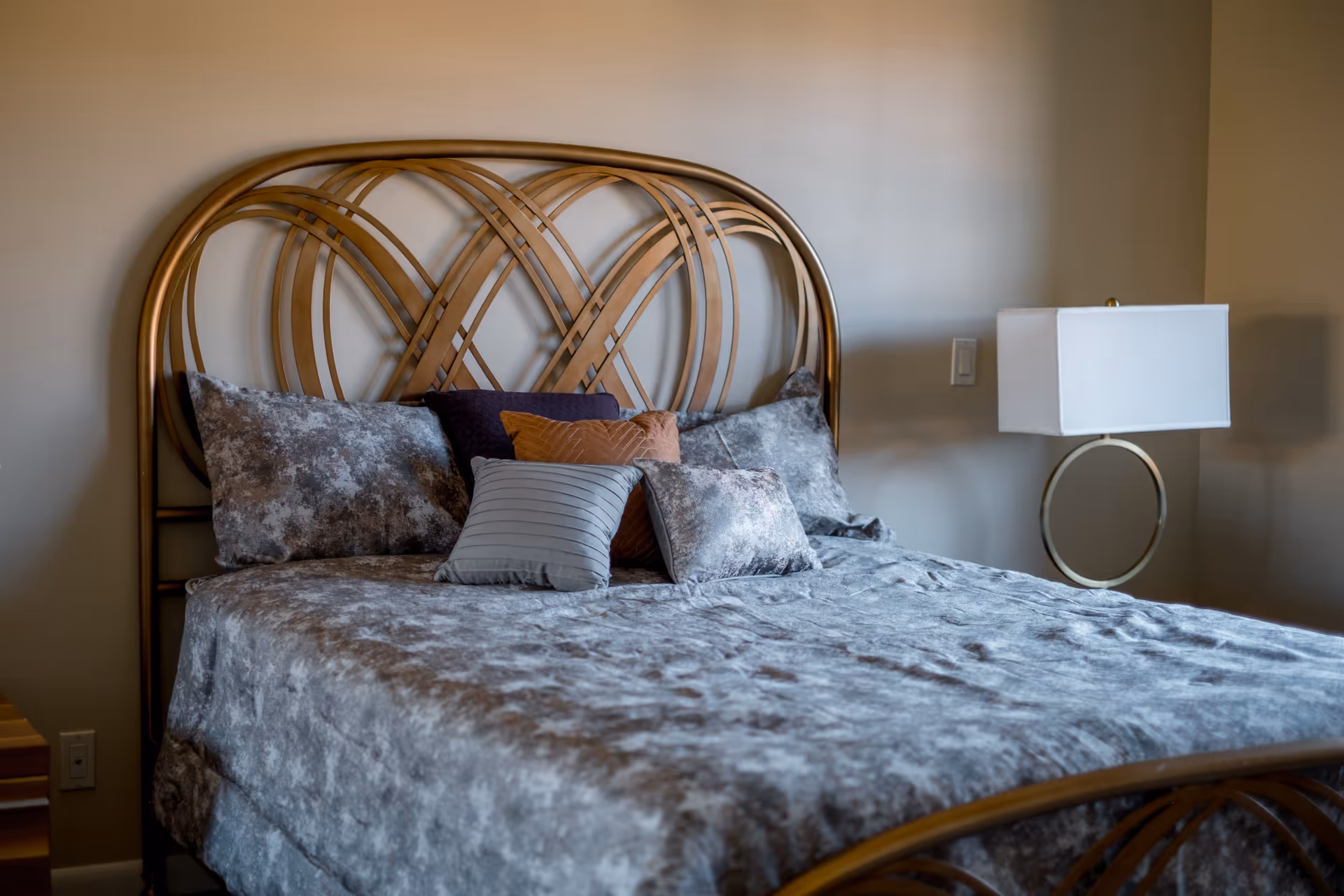 A neatly made bed with a decorative metal headboard, plush gray bedding and assorted pillows next to a bedside lamp.