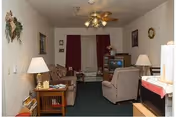 A cozy living room with a ceiling fan and light fixture, a patterned sofa, an armchair, a wooden side table with a lamp, and a TV on a stand. The room has dark green carpet, white walls decorated with framed pictures and a wall hanging, and a window with red curtains.