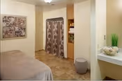 A cozy room with a massage or treatment table covered in a beige sheet, a decorative wall art piece above the table, a curtain covering a storage area, shelves with towels and supplies, a small round stool, and a countertop with a decorative bowl and a green plant.