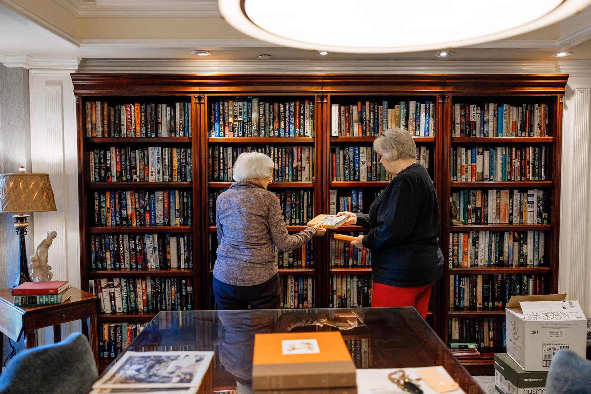 Two elderly women standing in front of a large wooden bookshelf filled with books in a cozy room. One woman is handing a book to the other. The room has a table with some items on it, a lamp on a side table, and a box on the floor.