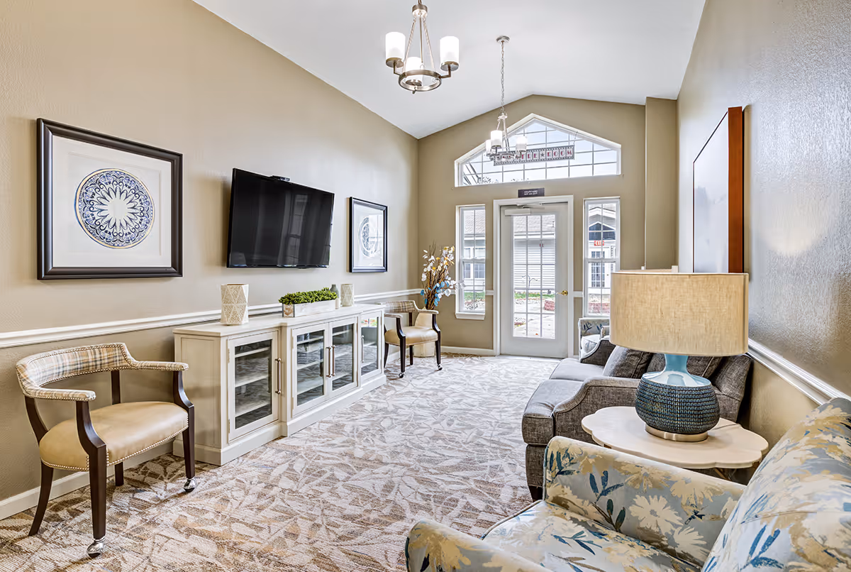 Bright common living room with sofas and chairs, a wall-mounted TV above a cabinet, decorative artwork, and a glass door with transom windows.
