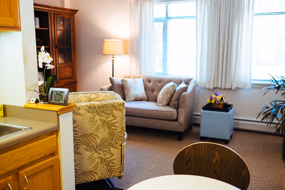 A bright living room with a sofa, patterned armchair, floor lamp, small table with flowers, and a kitchenette counter in the foreground.