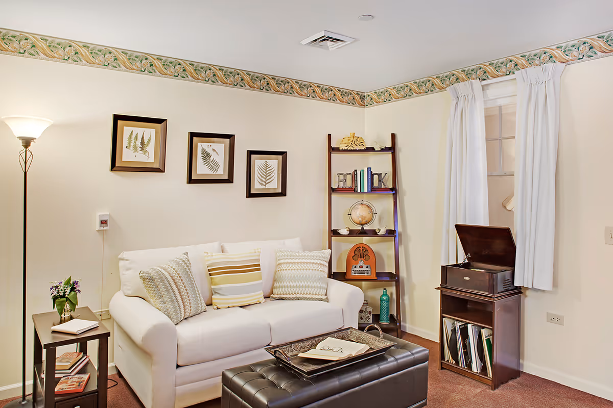 Cozy living room with a white sofa, patterned throw pillows, a floor lamp, bookshelf with decorative items, and a record player cabinet.