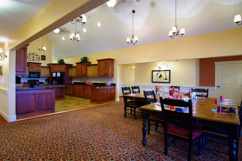 Interior view of a senior living facility dining area and kitchen. The dining area has wooden tables and black chairs with red cushions, decorated with small flower arrangements and place settings. The kitchen features wooden cabinets, black appliances, and a tiled floor. The ceiling is vaulted with multiple hanging light fixtures.