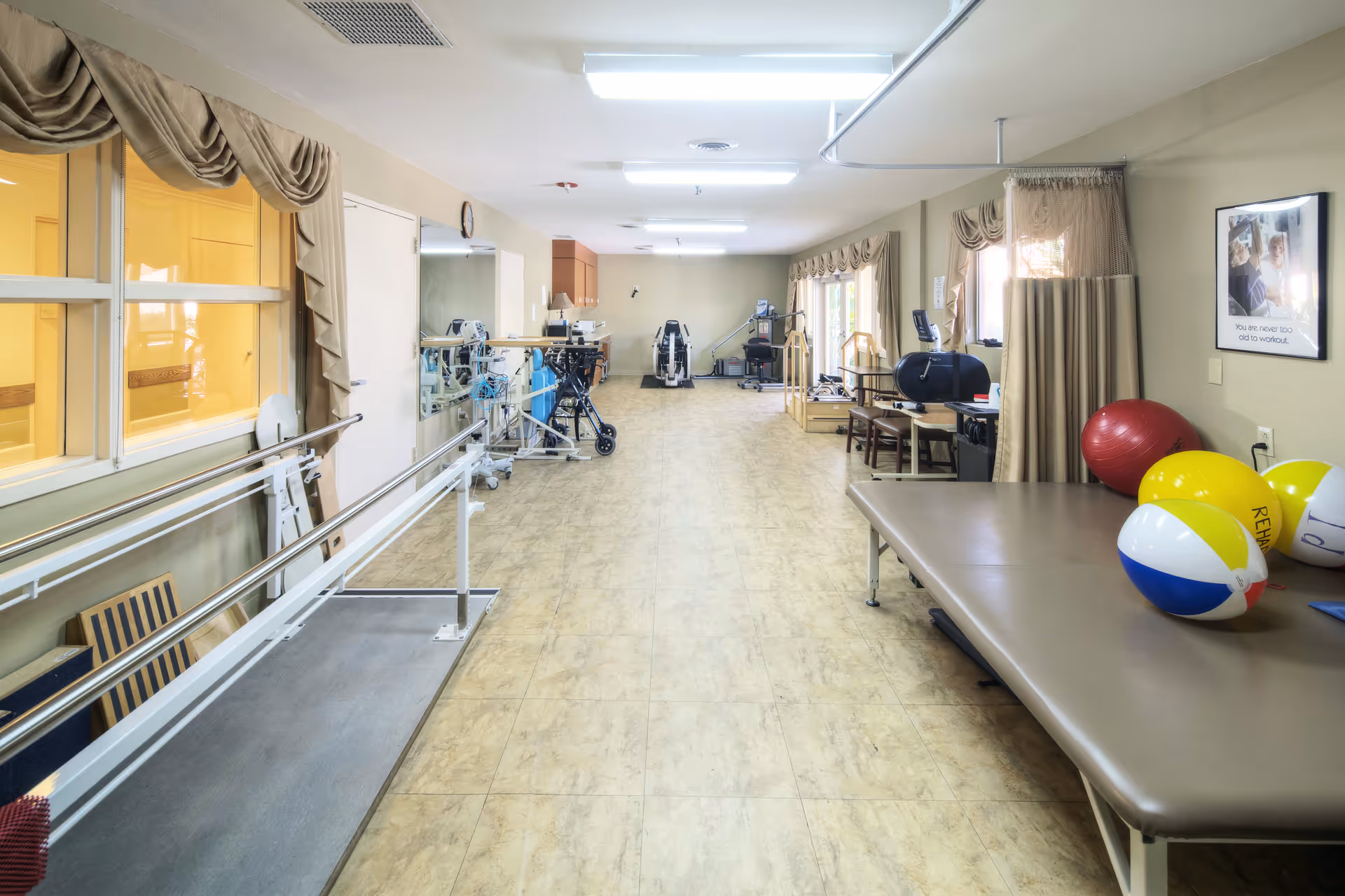 Long physical therapy room with parallel bars, treatment table, exercise equipment and colorful therapy balls.