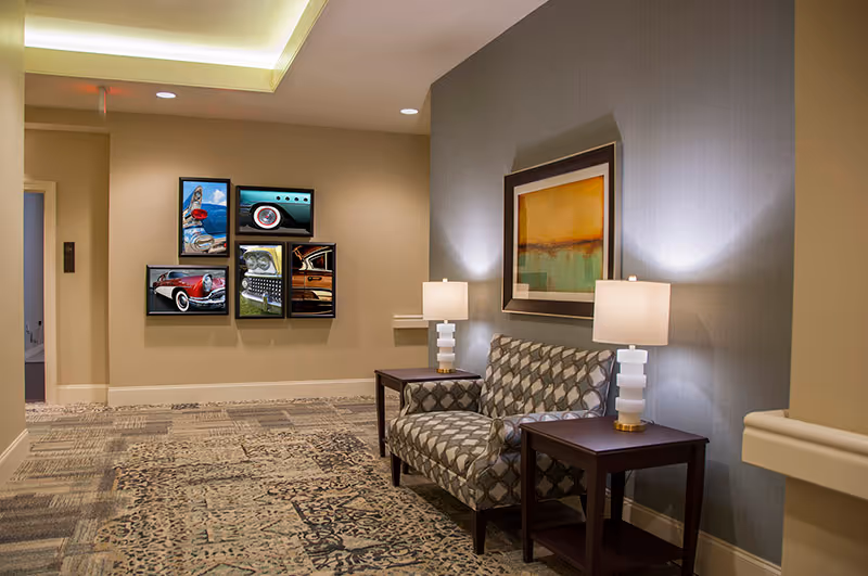A furnished hallway seating area with patterned armchairs, side tables and lamps, framed artwork, and a wall-mounted multi-panel picture.