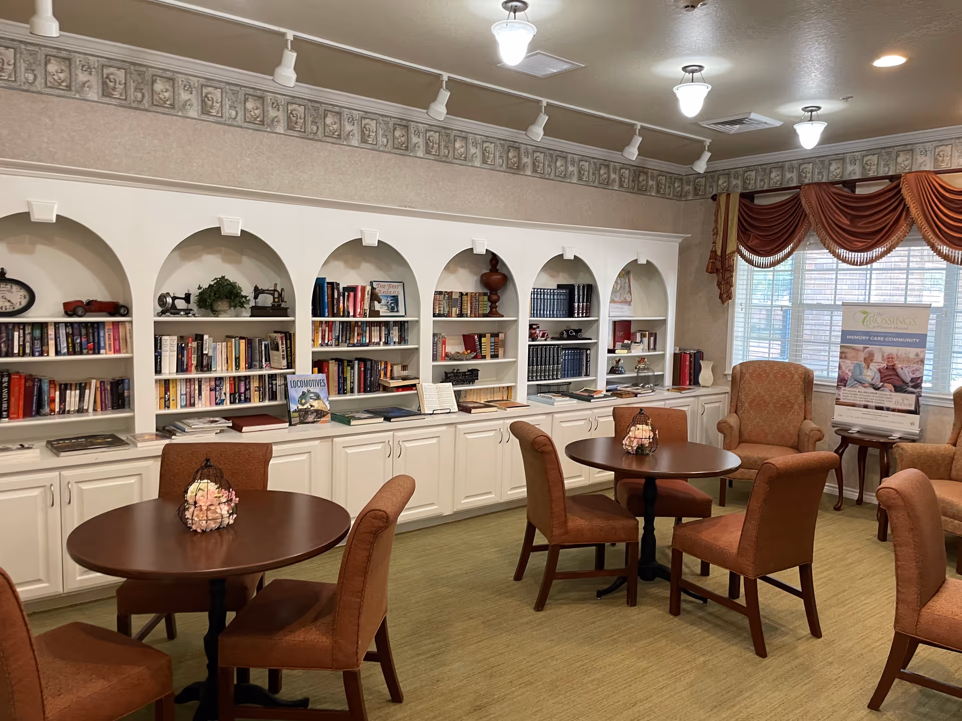 A cozy common area in an assisted living facility featuring round wooden tables with brown upholstered chairs and armchairs. The back wall has built-in white shelves with books, decorative items, and plants. Large windows with red drapes allow natural light to fill the room.