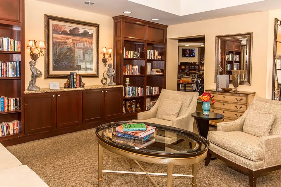 A cozy living room area in a retirement community featuring two beige armchairs with cushions, a glass-top oval coffee table with books on it, built-in wooden bookshelves filled with books, decorative statues, a framed landscape painting, wall sconces with lights, a wooden chest of drawers with a large mirror above it, and a small side table with a vase of flowers.