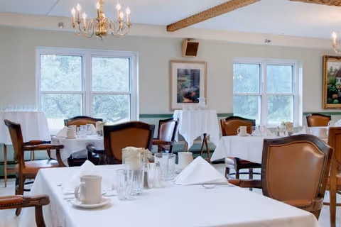 A bright dining room with several tables covered in white tablecloths, set with cups, glasses, napkins, and silverware. The room has large windows letting in natural light, wooden chairs with cushioned seats, chandeliers hanging from the ceiling, and framed artwork on the walls.