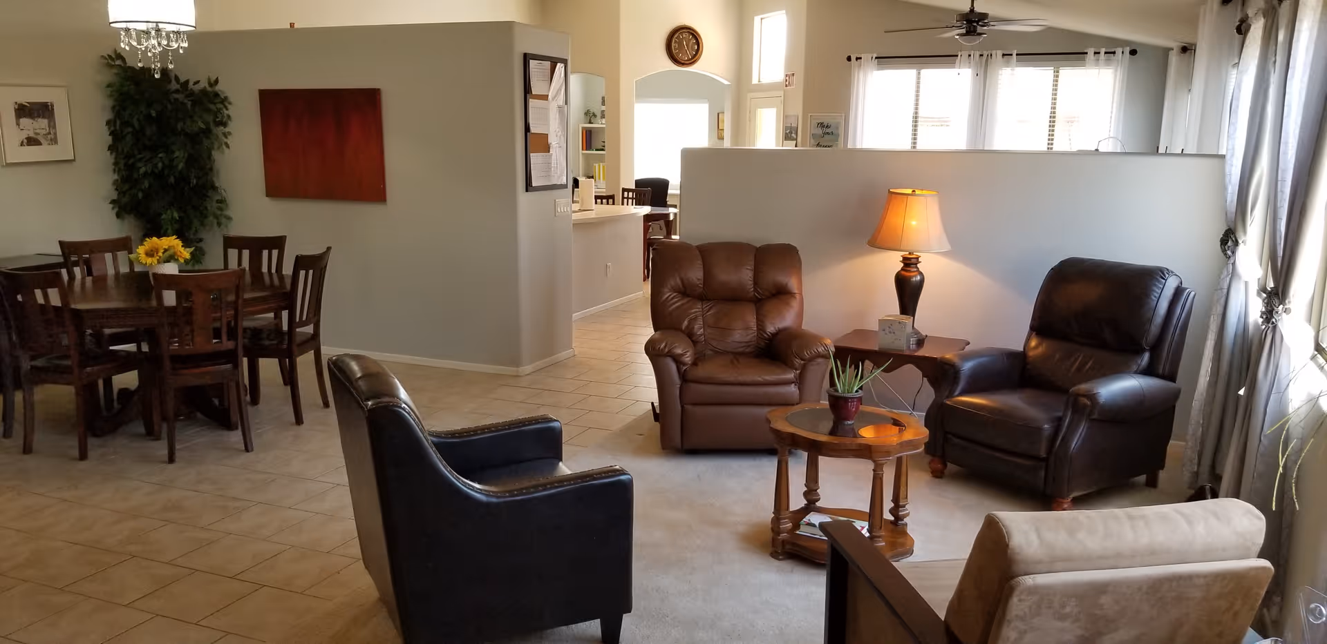 A cozy living room area with four armchairs arranged around a small wooden coffee table with a plant on it. A side table with a lamp and tissue box is next to one of the armchairs. In the background, there is a dining area with a wooden dining table and six chairs, a potted plant, and a red wall decoration. The room has tiled flooring and large windows with curtains letting in natural light.