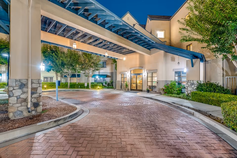 Entrance of Westminster Terrace Senior Living at night showing a covered driveway with hanging lanterns, stone pillars, and well-maintained landscaping including trees and bushes.