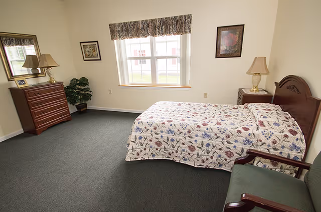 Furnished bedroom with a floral bedspread, wooden headboard, nightstand and lamp, dresser with mirror, chair, and a window.
