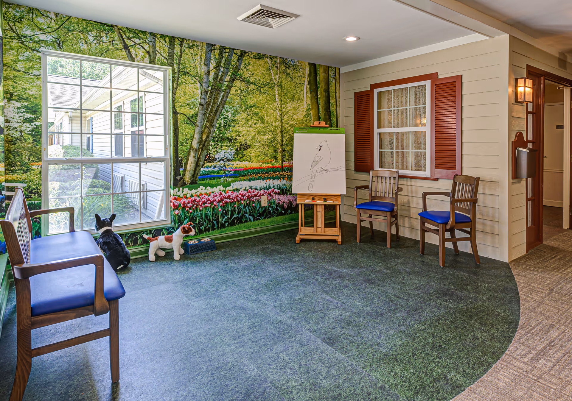 A cozy indoor seating area with wooden chairs featuring blue cushions, a bench, and two stuffed dog toys on the floor. The wall has a large mural depicting a vibrant garden with trees and colorful flowers. There is a window with a view of the outside building and an easel with a drawing of a bird on a branch.