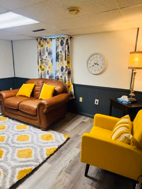 Cozy resident lounge with a brown leather sofa and a yellow chair, floral curtains, a wall clock, a side table with a lamp, and a patterned rug.