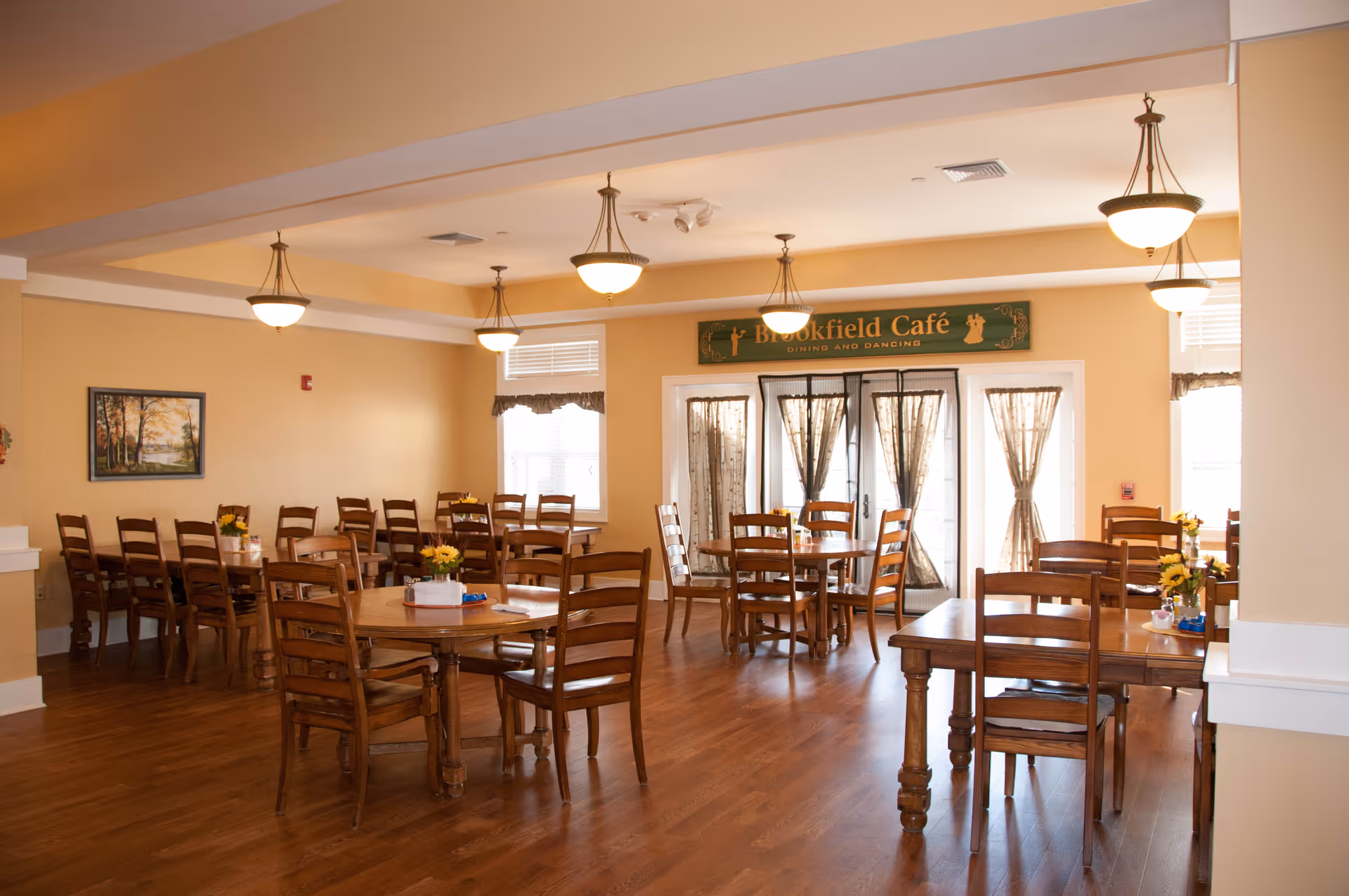 A dining room with wooden tables and chairs arranged neatly on a wooden floor. The room has warm beige walls and multiple hanging light fixtures. There are windows with curtains letting in natural light, and a sign above a set of glass doors reads 'Brookfield Café Dining and Dancing'. Small flower arrangements are placed on the tables.