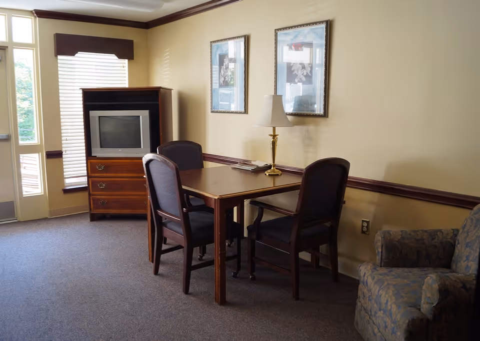 A small sitting area with a wooden table and four chairs, a table lamp, two framed pictures on the wall, a TV on a wooden cabinet, and a patterned armchair. There is a window with blinds and a door letting in natural light.
