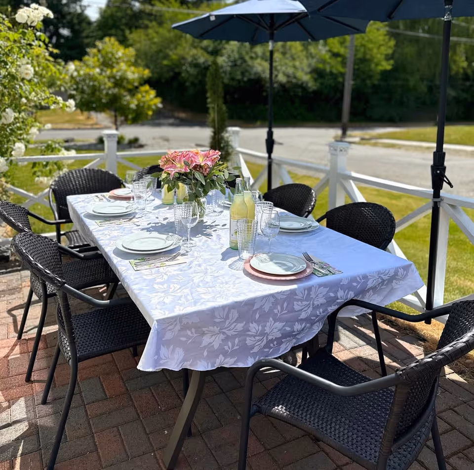 Outdoor patio dining area with a rectangular table covered with a white floral tablecloth, set with plates, glasses, cutlery, and bottles of lemonade. There are six black wicker chairs around the table and a blue umbrella providing shade. The background shows greenery and a white fence.
