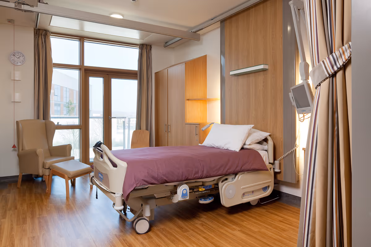 A clean and modern hospital-style bedroom with a single adjustable bed covered with a purple blanket and white pillows. The room features wooden flooring, a beige armchair with a matching footrest, a wooden chair, large windows with beige curtains, and wooden cabinets. A medical monitor is mounted on the wall next to the bed.