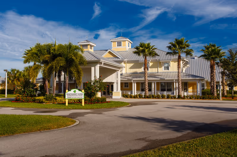 Exterior view of a senior living facility building with a light yellow facade, white trim, and a metal roof. The building is surrounded by palm trees and well-maintained landscaping under a partly cloudy blue sky. A sign in front reads 'Independent Living Administration & Sales'.