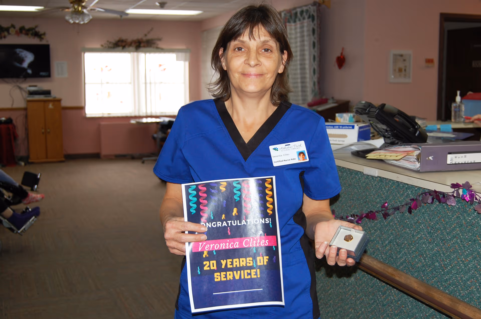 A woman in blue scrubs stands inside a facility holding a certificate that reads 'Congratulations! Veronica Clites 20 Years of Service!' and a small box with a pin. She is smiling and wearing a name badge. The background shows an office or reception area with a counter, phone, and some decorations.
