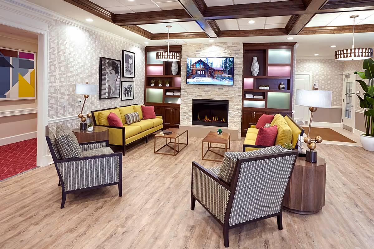 Bright assisted-living common room with sofas and armchairs arranged around a fireplace and wall-mounted TV.