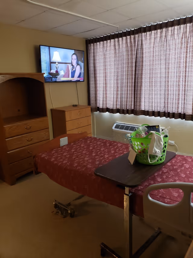 A senior living facility bedroom with a hospital-style bed covered in a red patterned blanket. A green basket with various items and a face mask is on an overbed table. There is a wooden dresser and cabinet, a wall-mounted TV showing a woman, and a window with closed brown curtains. An air conditioning unit is below the window.