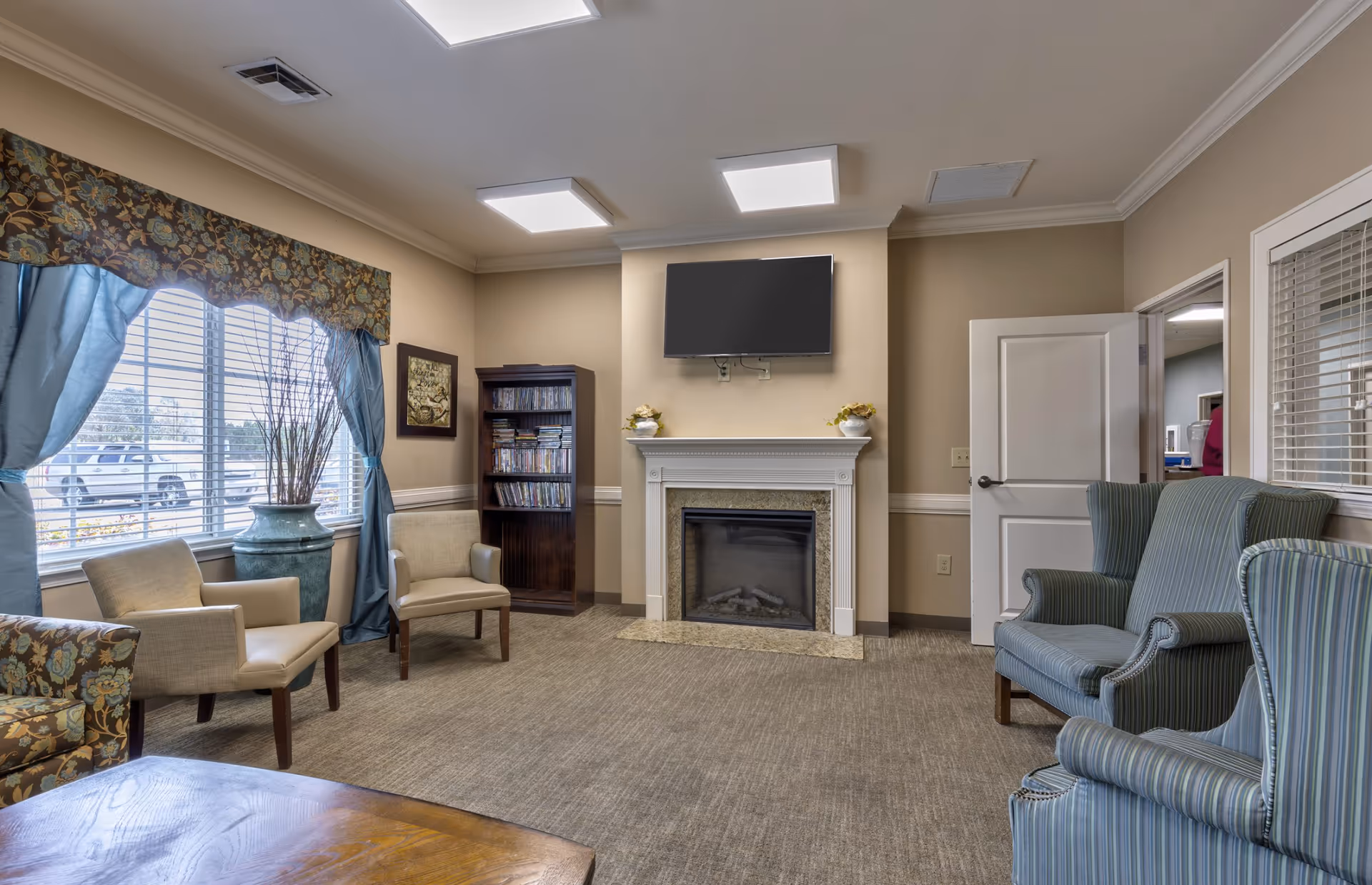 Cozy common room with chairs, a fireplace topped by a wall-mounted TV, bookshelf, and a large window with curtains.